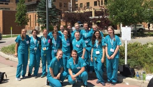 A group of students in blue scrubs are smiling to the camera in front of a hospital