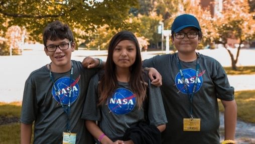3 students in NASA shirts are smiling at the camera, arms around one another.